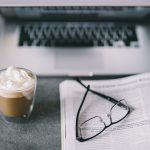 Close up of glasses, magazine, and coffee in front of a laptop
